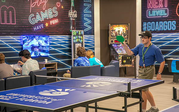 A student about to hit a ping pong ball at a ping pong table with a ping pong paddle in the UWF Game Room while four students are playing video games in the background.