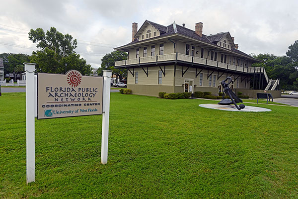 Two-story historic building with wraparound veranda on the second story. A large ship anchor and a Florida Public Archaeology Network Coordinating Center sign are displayed in front of the building.