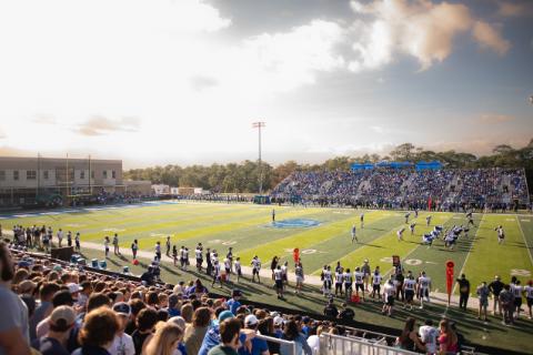 A UWF football game played on a football field watched from the visitors side bleachers point of view.